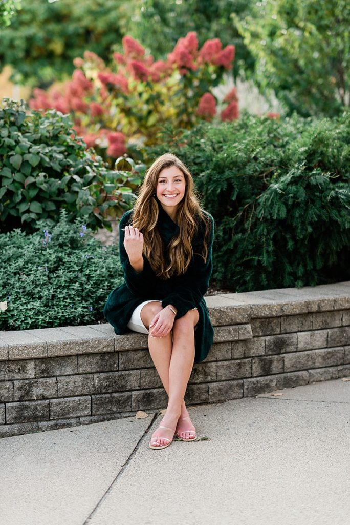 Ideas and locations for a Michigan State University fall senior photo session on north campus by Allie Siarto Photography, East Lansing Photographers. A photo of an MSU senior sitting on a stone wall with colorful flowers behind her.
