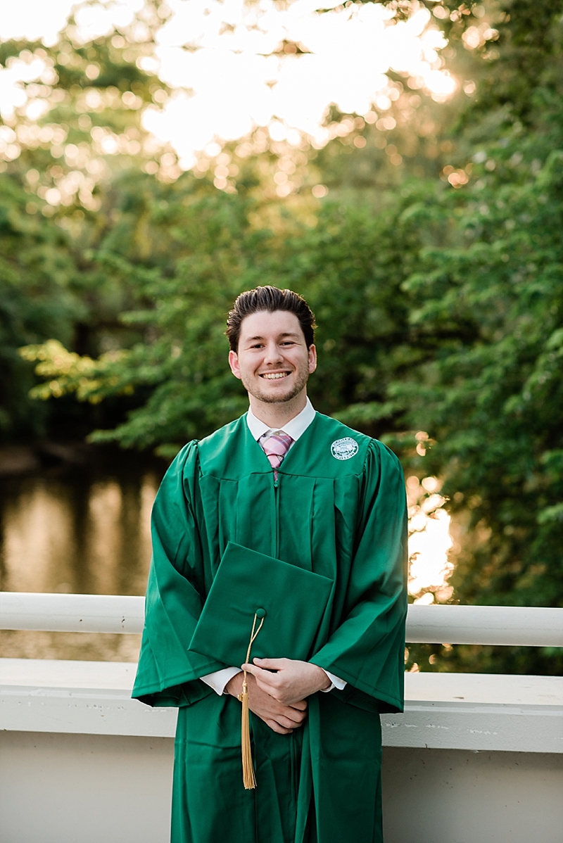 Michigan State Senior Pictures with a guy in a cap and gown on MSU's campus on the bridge by the library and Spartan Stadium overlooking the Red Cedar River, by Allie Siarto & Co. Photography, MSU graduation photographers