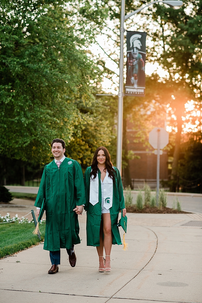 Michigan State Senior Pictures with a couple in a cap and gown walking on MSU's campus near the Sparty statue with the sunset in the background, by Allie Siarto & Co. Photography, MSU graduation photographers