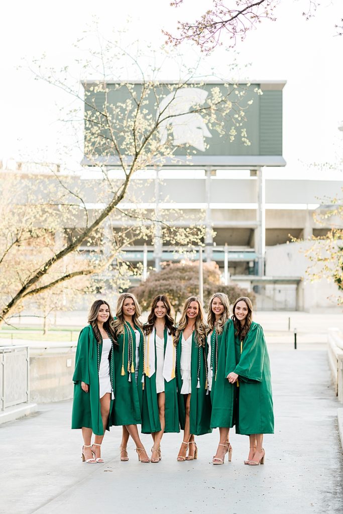 Michigan State University senior photography ideas with roommates on campus in white dresses and cap and gown on the bridge behind the library with the Spartan Stadium Sparty head in the background, by Allie Siarto & Co, senior photographers in East Lansing, Michigan