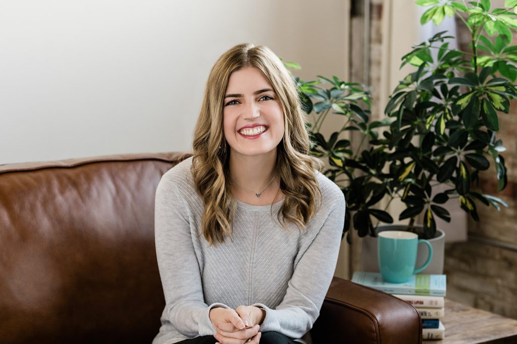 Lansing, Michigan headshot photographer with personal branding photo of a friendly therapist sitting on brown leather couch smiling at the camera with plants and a coffee cup in the background