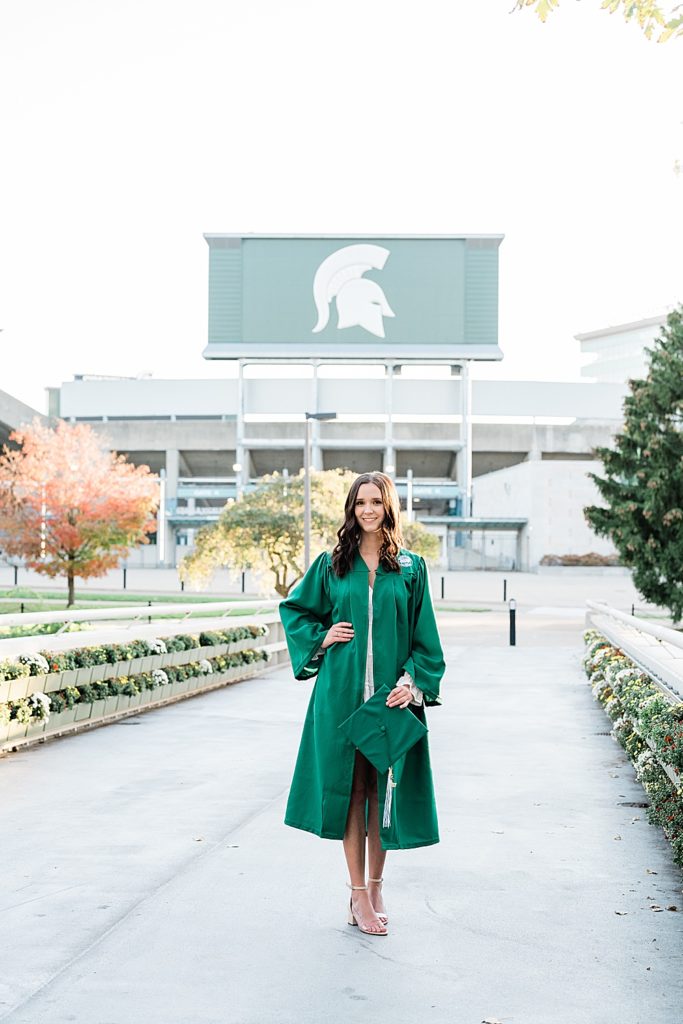 Michigan State graduation pictures on north campus in the fall with a white dress and grad gown holding a cap with Spartan Stadium in the background by Allie & Co. Photography, East Lansing photographers