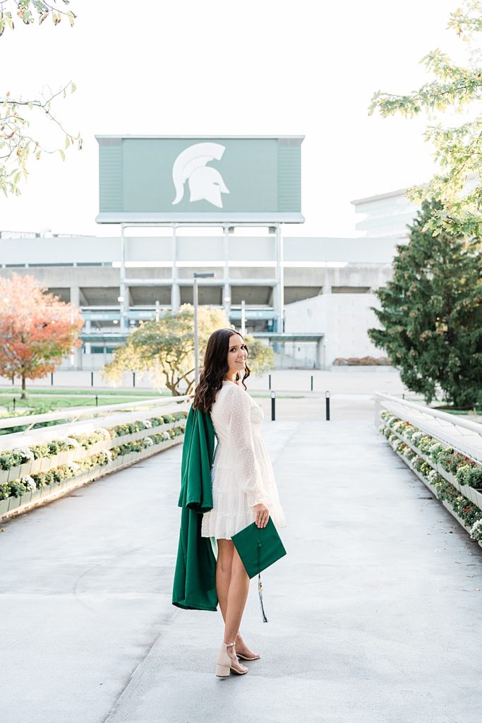 Michigan State graduation pictures on north campus in the fall with a white dress and grad gown over the shoulder with Spartan Stadium in the background by Allie & Co. Photography, East Lansing photographers