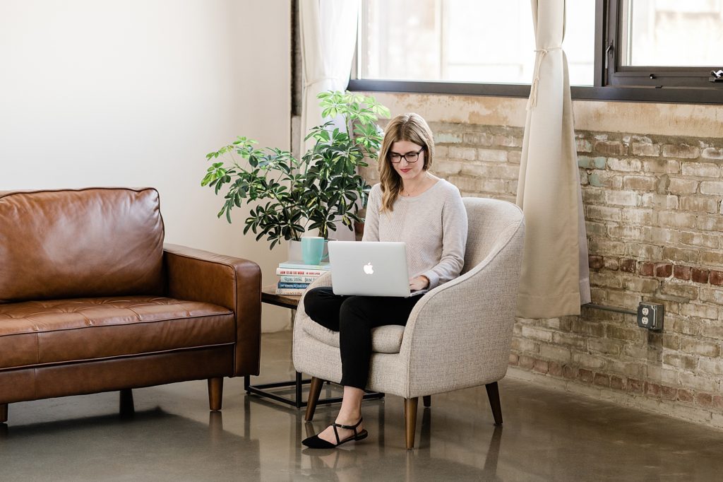 Lansing head shot photographer, photo of a woman sitting on a gray chair with a laptop in her lap, in a modern loft room with concrete floors, by Allie Siarto & Co. Photography, Michigan branding photographers