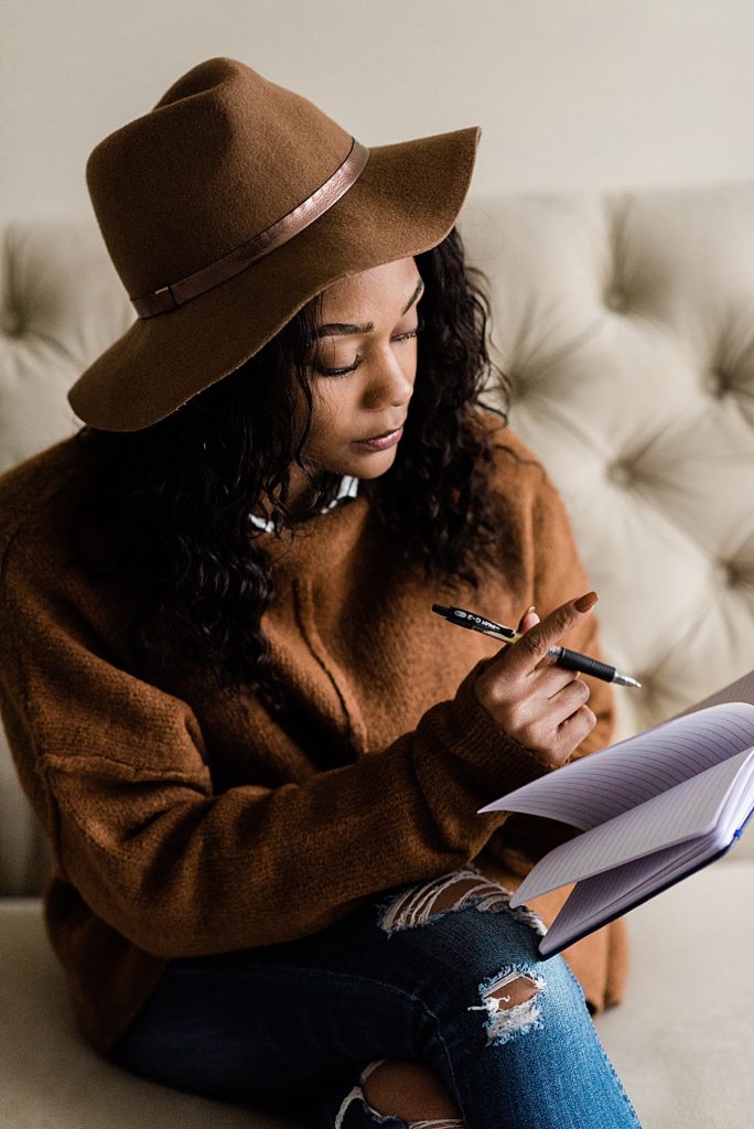 Lansing head shot photographer, photo of a woman sitting on a tufted tan couch with a felt hat on and writing in her notebook, by Allie Siarto & Co. Photography, Michigan branding photographers