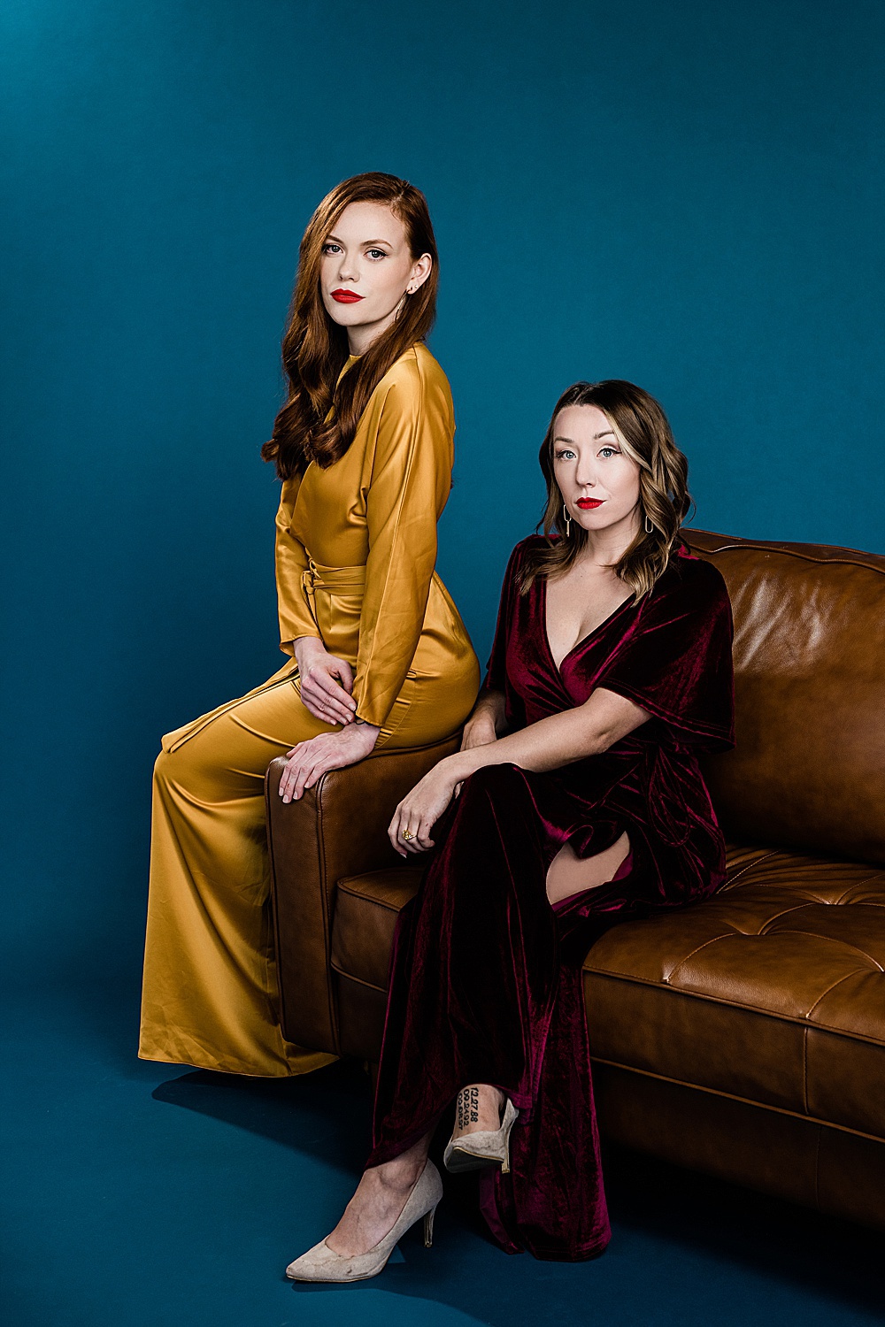 Lansing, Michigan commercial fashion photography, photo of two models - one in a yellow dress and one in a red velvet dress sitting on a brown couch on a seamless blue backdrop by Allie Siarto & Co., Lansing commercial photographer