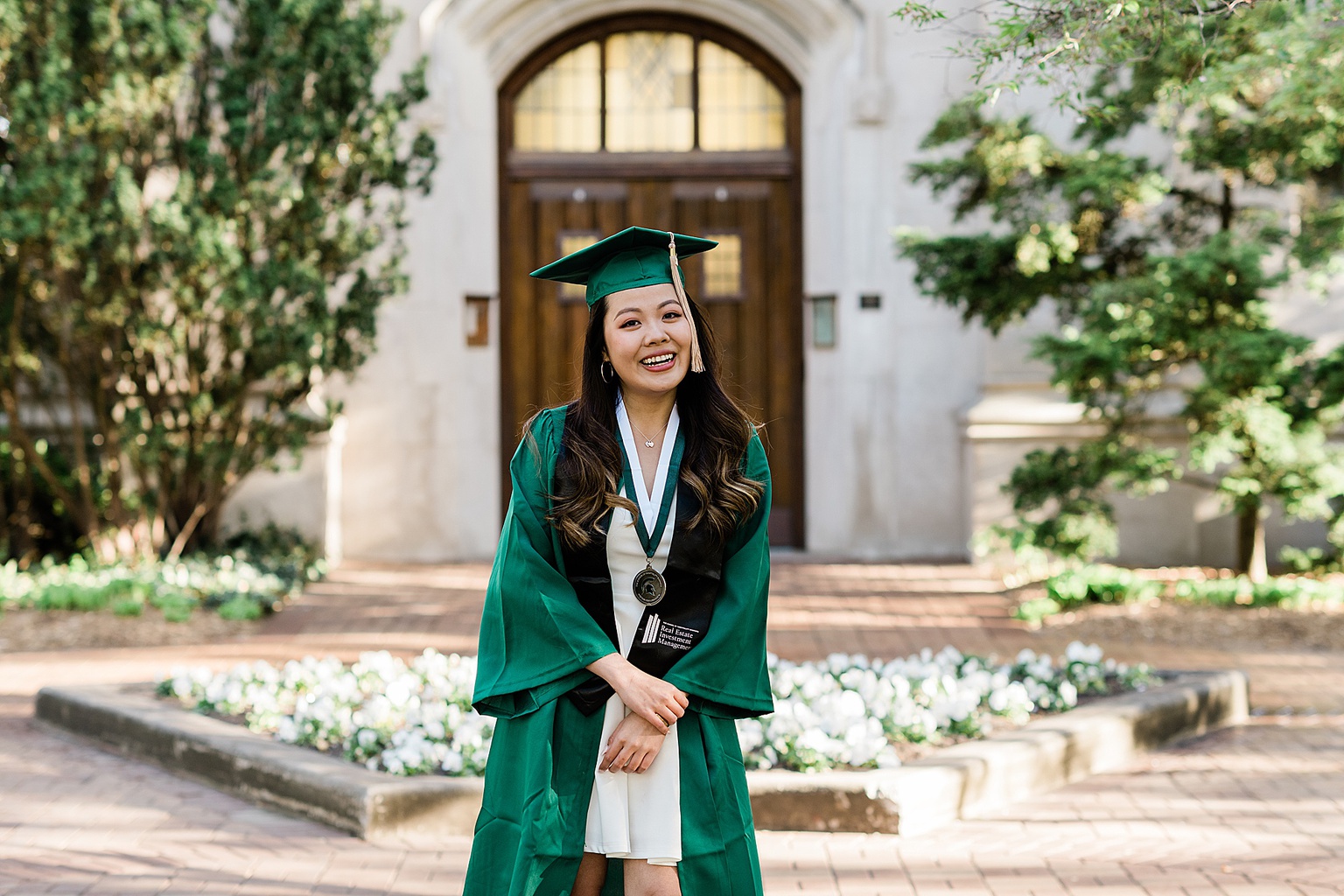 Michigan State Senior grad photoshoot on MSU's campus, photo of a woman in a white dress and cap and gown standing in front of the Beaumont Tower door, by Allie Siarto & Co., East Lansing photographers