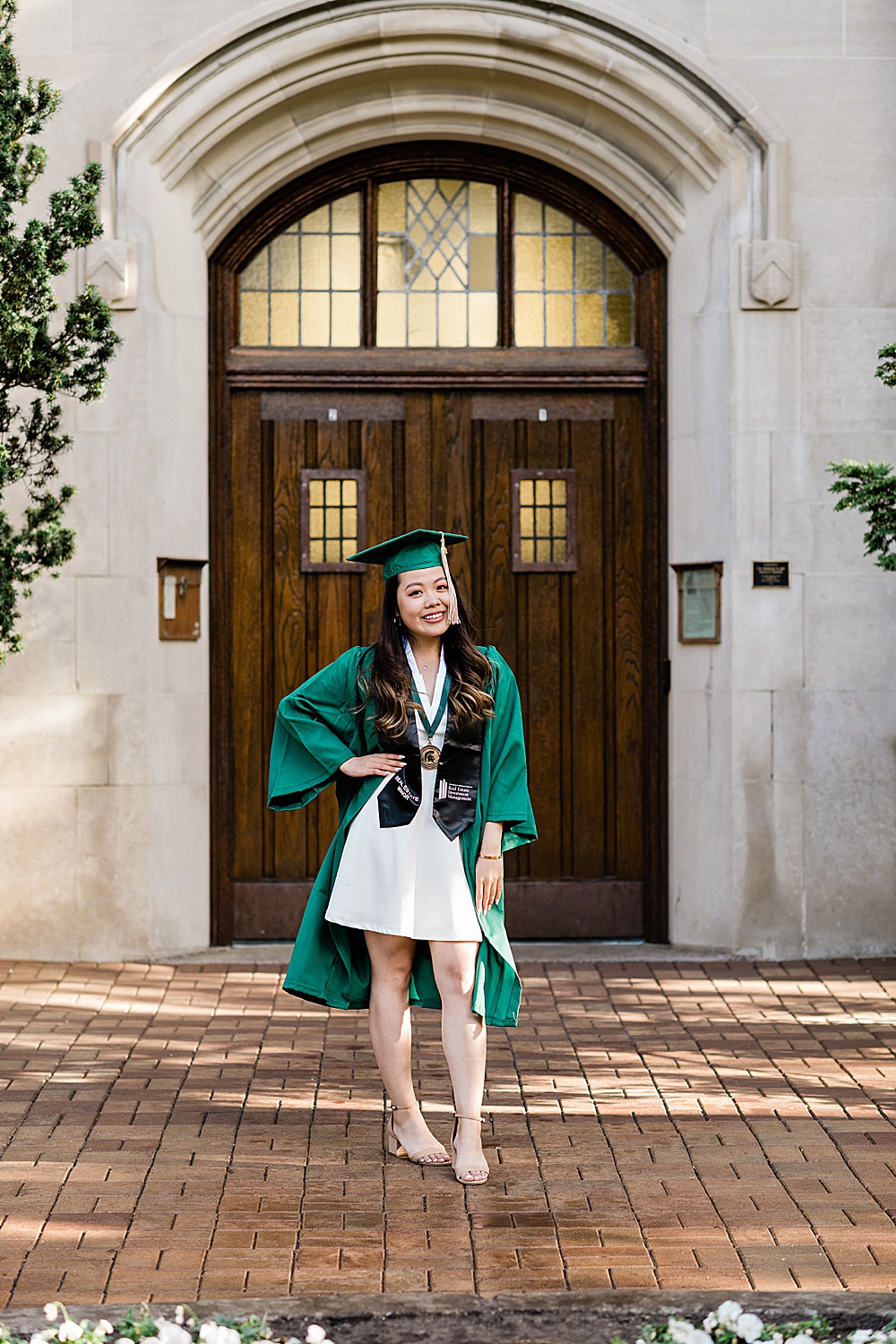 Michigan State Senior grad photoshoot on MSU's campus, photo of a woman in a white dress and cap and gown standing in front of the Beaumont Tower door, by Allie Siarto & Co., East Lansing photographers