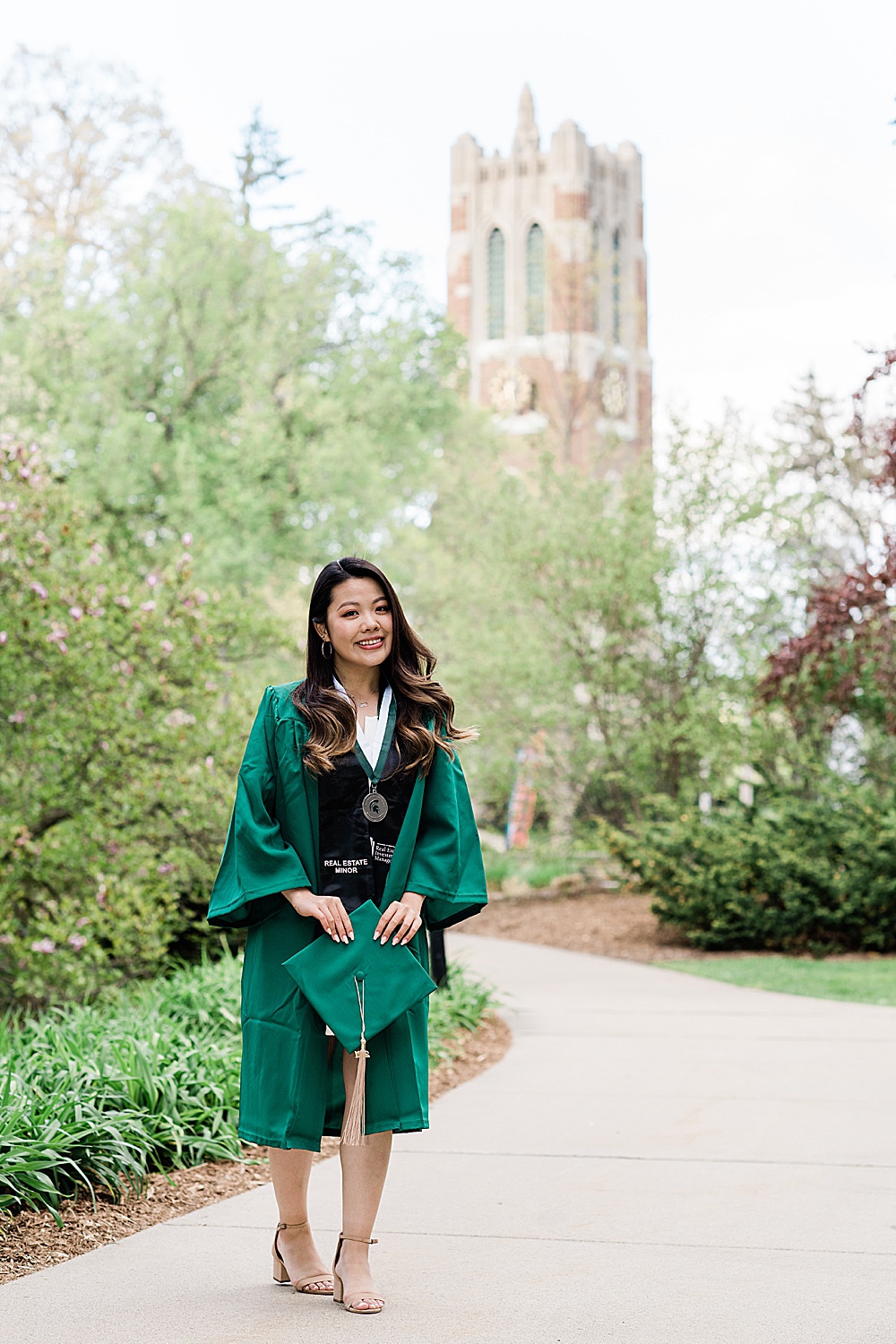 Michigan State Senior grad photoshoot on MSU's campus, photo of a woman in a graduation gown holding her grad cap with Beaumont Tower behind her, by Allie Siarto & Co., East Lansing photographers