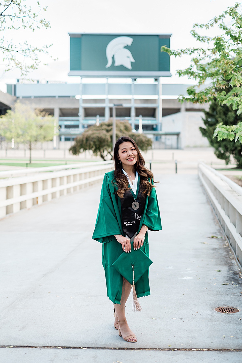 Michigan State Senior grad photoshoot on MSU's campus, photo of a woman in a white dress wearing graduation gown on the bridge by Spartan Stadium, by Allie Siarto & Co., East Lansing photographers