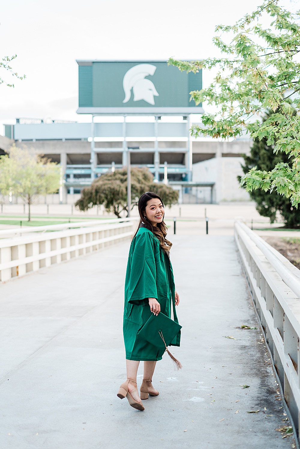 Michigan State Senior grad photoshoot on MSU's campus, photo of a woman in a white dress wearing graduation gown on the bridge by Spartan Stadium, by Allie Siarto & Co., East Lansing photographers