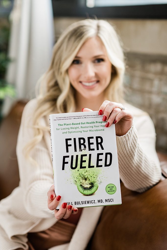 Nurse injector branding photos, lounging on a brown leather couch in loungewear and holding out a book title "Fiber Fueled" with a brick wall and plants in the background, by Allie Siarto & Co., Michigan headshot and branding photographers