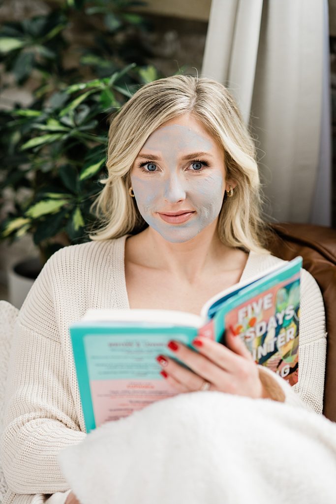 Nurse injector branding photos, lounging on a brown leather couch in loungewear and reading a book with a face mask on with a brick wall and plants in the background, by Allie Siarto & Co., Michigan headshot and branding photographers