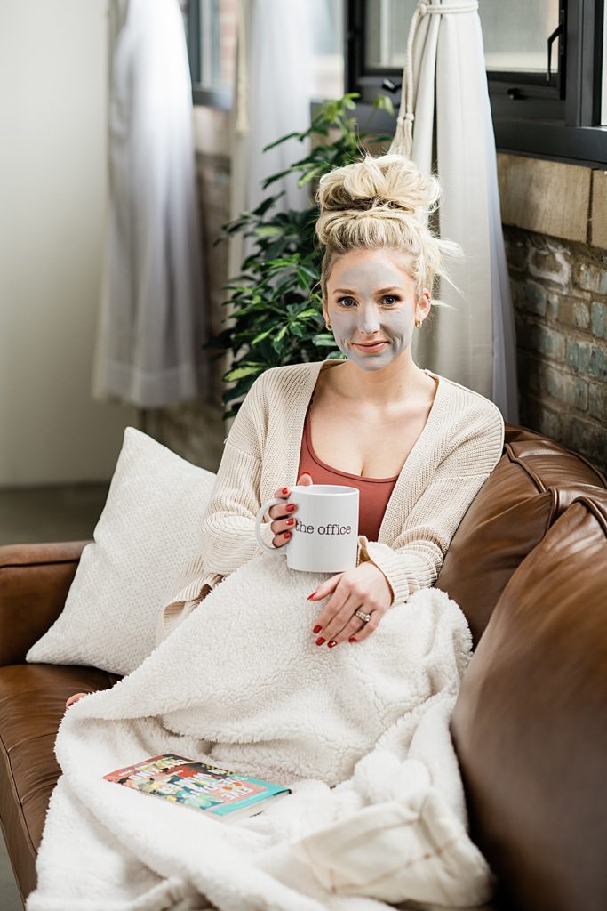 Nurse injector branding photos, lounging on a brown leather couch under a white blanket in loungewear and top bun, holding a coffee mug with a face mask on, with a brick wall and plants in the background, by Allie Siarto & Co., Michigan headshot and branding photographers