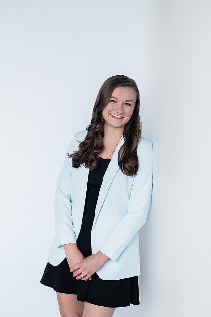 Lansing, Michigan headshot branding photos of a young photographer leaning against a white wall with a light blue jacket and black dress