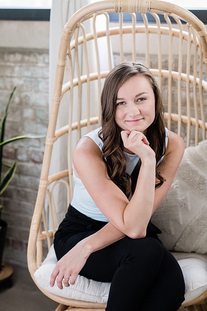 Lansing, Michigan headshot branding photos of a young photographer sitting on a wooden boho chair with a plant and brick wall behind her