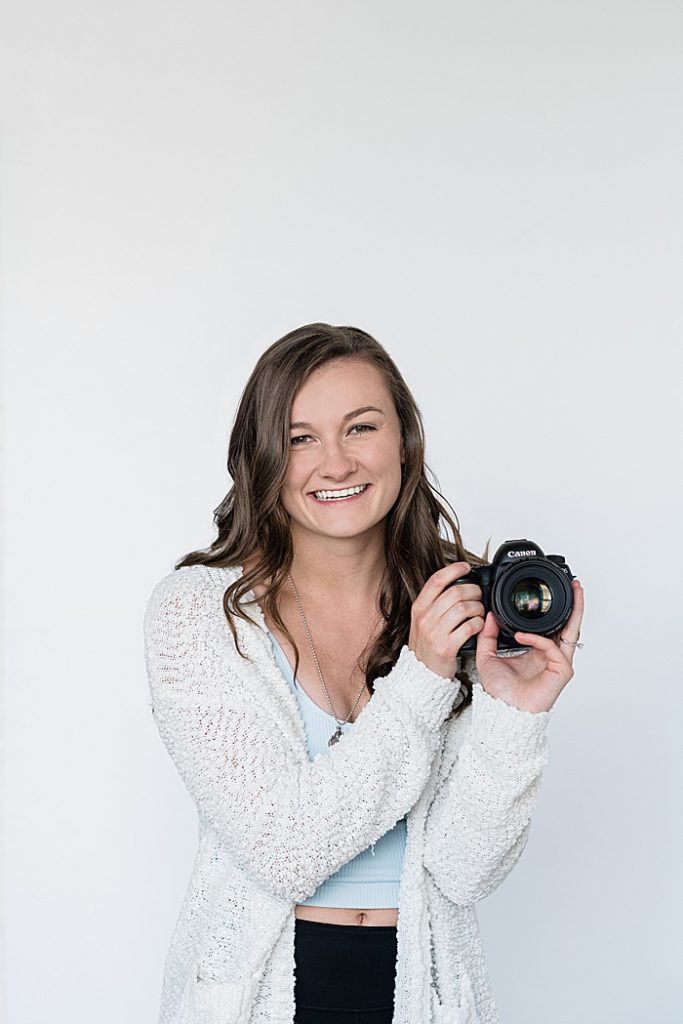 Lansing, Michigan headshot branding photos of a young photographer against a white backdrop holding her camera, wearing a blue shirt and white sweater