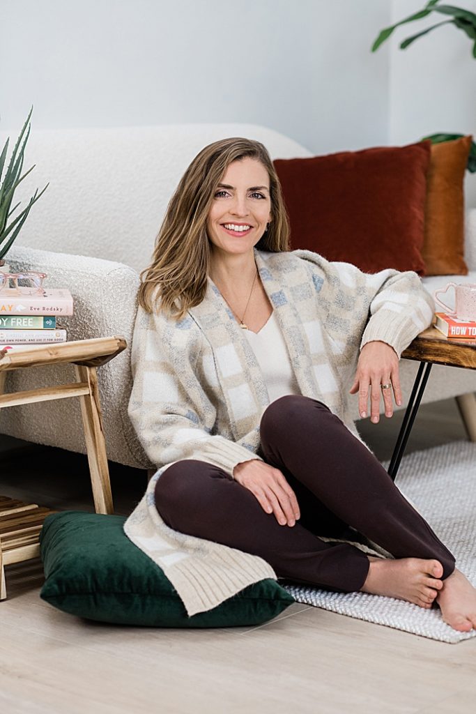 Branding headshot photo of a woman sitting on a light wooden floor leaning back against a white couch and a wooden coffee table surrounded by books, plants, and pillows, by Allie Siarto & Co. Photography, Lansing, Michigan headshot photographers
