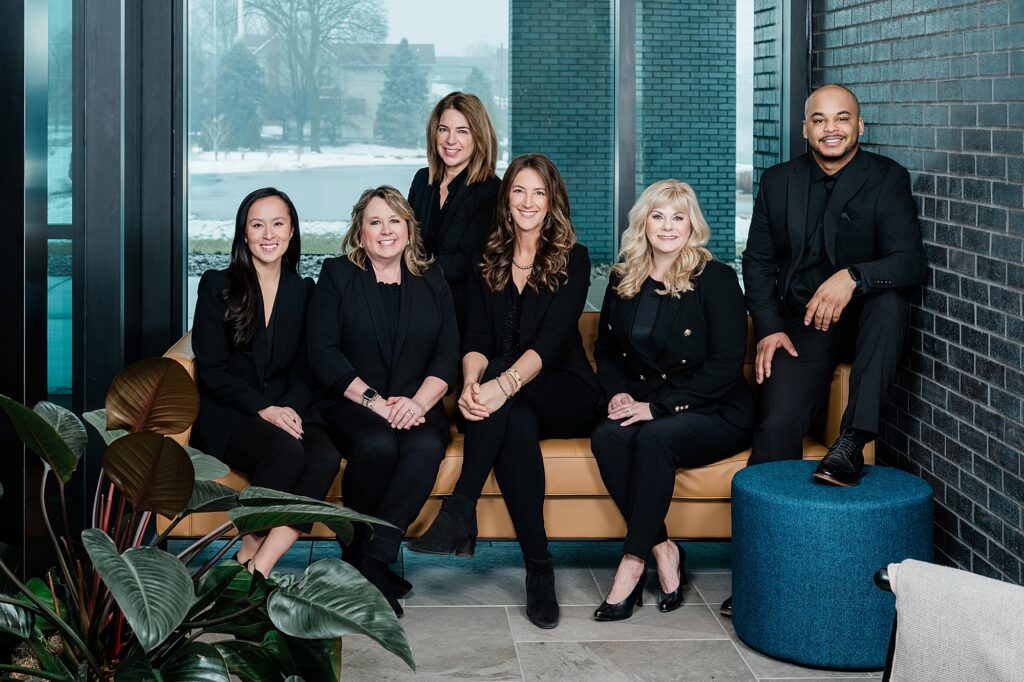 Marketing photos for a team of Lansing realtors, by Allie Siarto Photography, Lansing headshot and branding photographer; a realtor team photo with clean, modern background, including brick, glass, metal, and a plant in the background in a natural indoor setting
