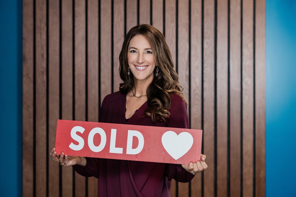 Marketing photos for a team of Lansing realtors, by Allie Siarto Photography, Lansing headshot and branding photographer; a photo of a realtor on a wood background holding a "sold" sign