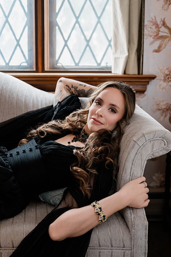 Creative business headshots: Audio book narrator Bridgerton inspired headshots by Allie Siarto Photography, headshot and portrait photographer out of East Lansing, Michigan - a woman lays on a vintage couch in a black dress with tattoos