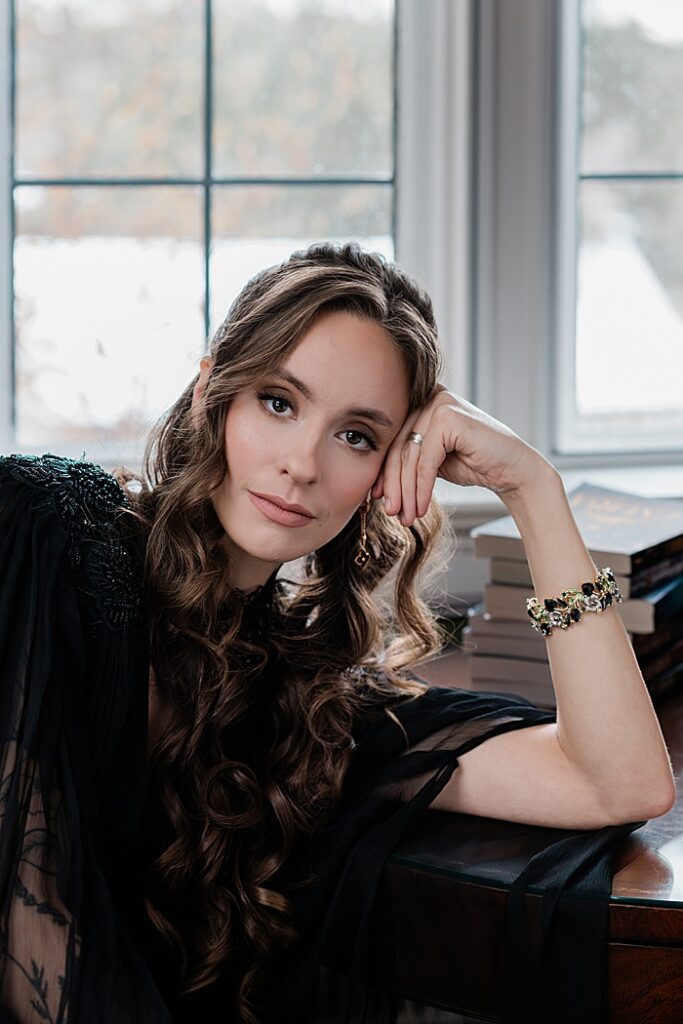 Michigan creative business photos: Audio book narrator Bridgerton inspired headshots by Allie Siarto Photography, headshot and portrait photographer out of East Lansing, Michigan - an audiobook narrator reads leans on a desk with books behind her in a Victorian setting