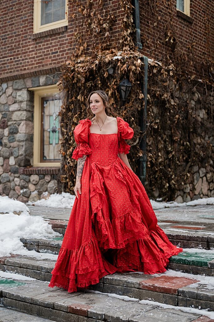 corporate photographer Michigan: Audio book narrator Bridgerton inspired headshots by Allie Siarto Photography, headshot and portrait photographer out of East Lansing, Michigan - an audiobook narrator holds out her red Victorian dress on the steps outside in a snowy setting