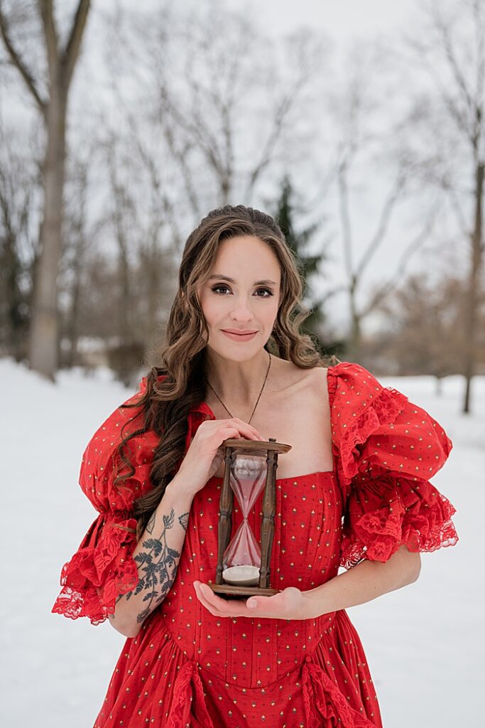 corporate photographer Michigan: Audio book narrator Bridgerton inspired headshots by Allie Siarto Photography, headshot and portrait photographer out of East Lansing, Michigan - an audiobook narrator holds an hourglass in a red Victorian dress outside in a snowy setting